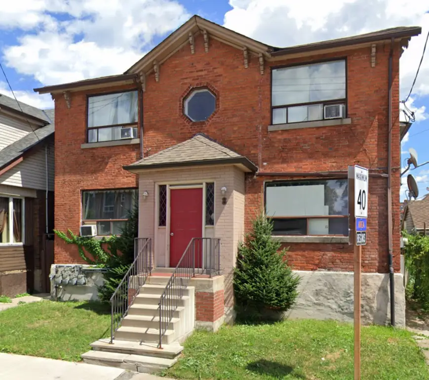 A two-story brick house with a gabled roof, a central recessed entrance flanked by stairs, and a circular window above the entrance. The house features large square windows with air conditioning units. The front yard has green grass and a small bush, showcasing prime Hamilton property management.