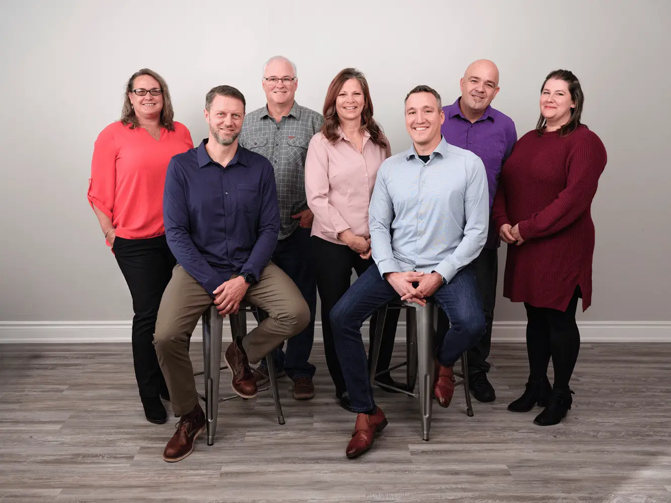A group of seven people—three women and four men—pose for a photo against a plain light-colored backdrop. Two men are seated on stools while the rest stand around them. They are dressed in business casual attire, representing Hanlyn Property Management, and are smiling at the camera.