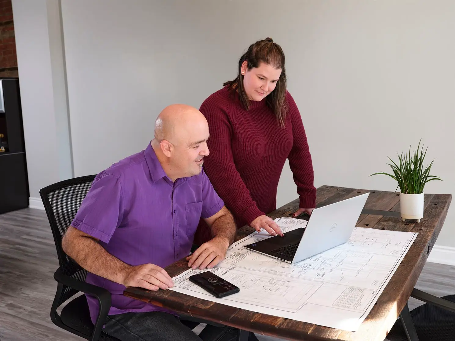Two people are collaborating at a wooden table. One individual, seated in a purple shirt, is examining blueprints while using a laptop, likely for property management services. The other person, standing in a maroon sweater, points at something on the screen. A smartphone and a potted plant are also on the table.